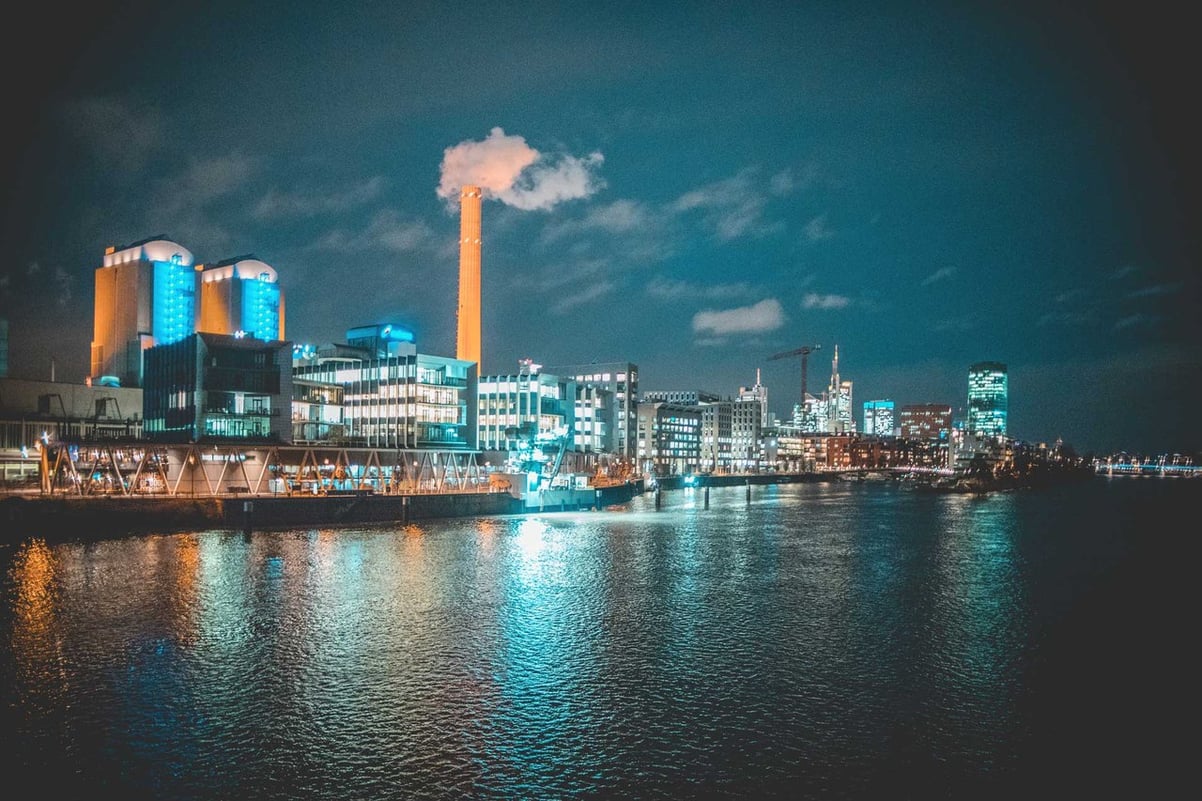 Nachtansicht einer beleuchteten Industrieanlage am Wasser mit rauchendem Schornstein